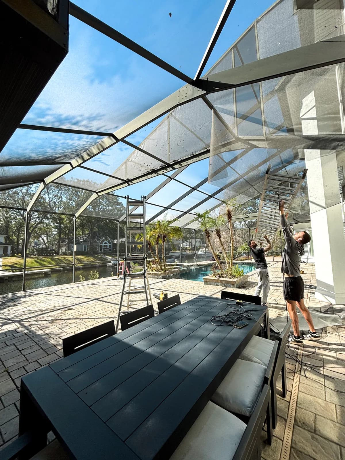 Two Screen'D In crew members rescreening pool enclosure roof panels from ladders